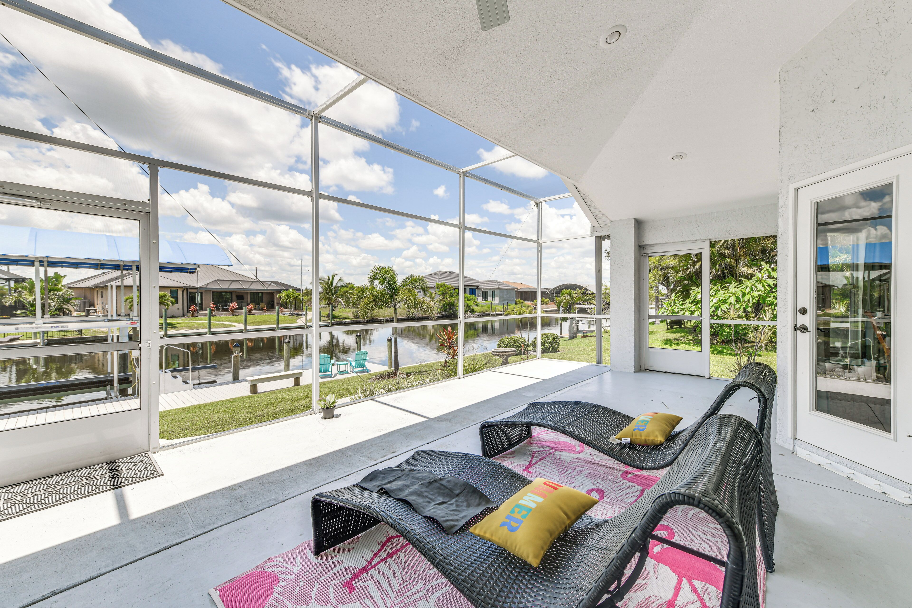 A covered outdoor seating area with wicker furniture and cushions, large glass windows offering views of a lake and surrounding greenery.