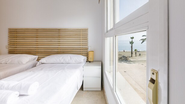 A bedroom with two beds, a nightstand, a lamp, and a view of the beach.