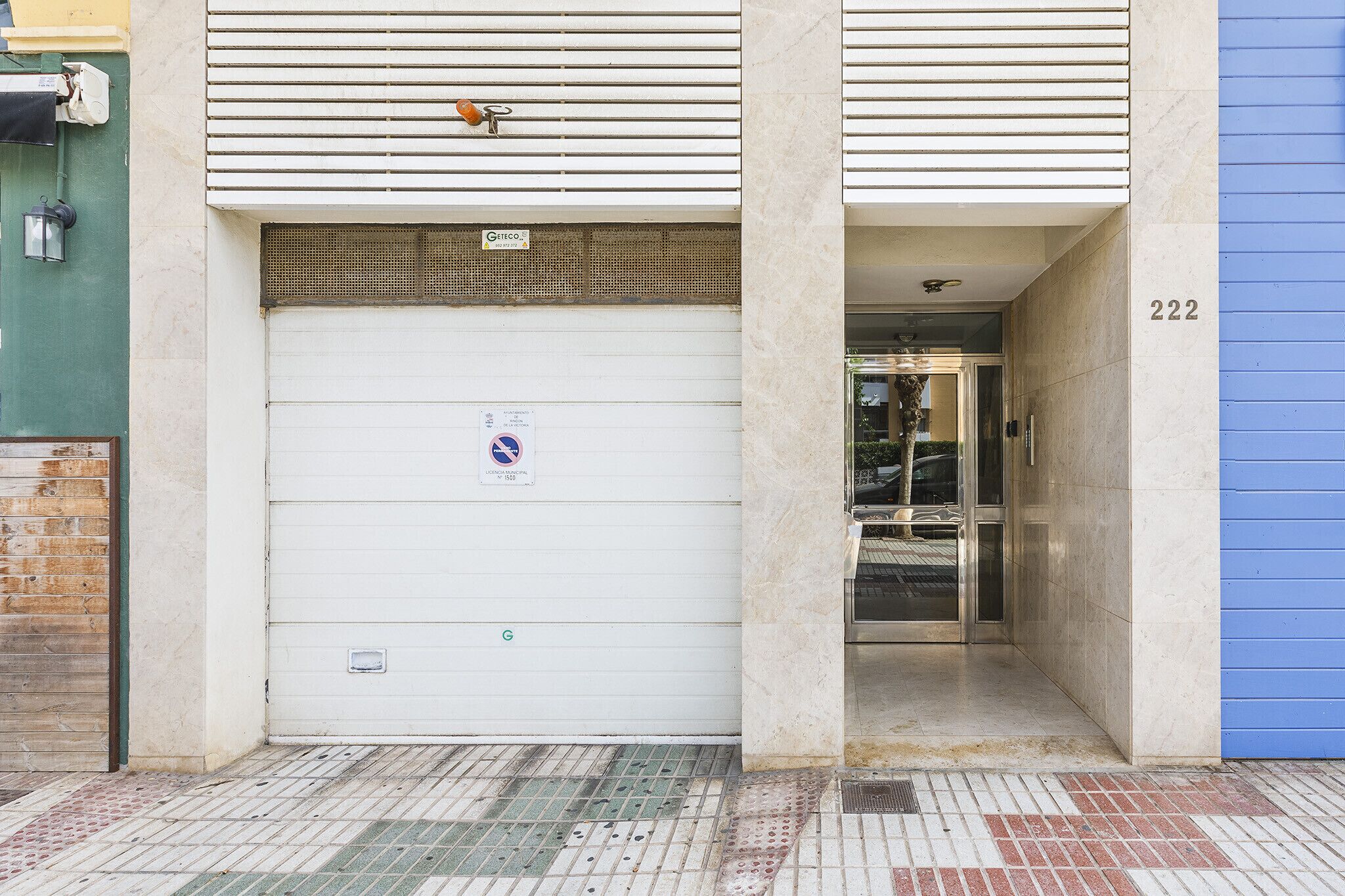 A building entrance with a closed garage door, a visible door number '222', and a tiled ground with a tactile paving strip.