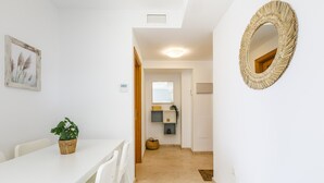 A white dining area with a table and chairs, a potted plant, and a woven mirror on the wall.