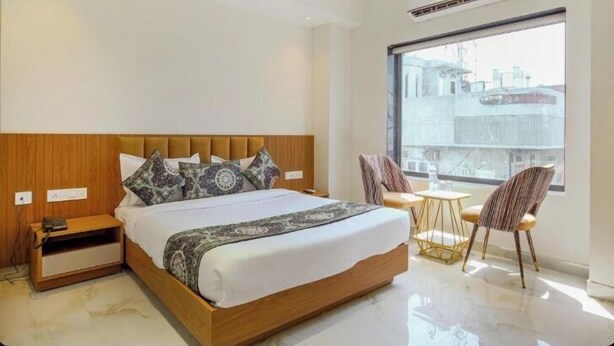 Una habitación de hotel moderna con una cama grande, un cabecero de madera, una mesita de noche con un teléfono y una pequeña mesa con sillas junto a un ventanal amplio.