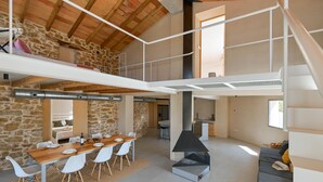 A spacious loft-style room with a high ceiling, exposed wooden beams, a stone wall, a dining area with a long wooden table and white chairs, a black chimney, and a grey sofa. - La de Ros - Javea, Costa Blanca (Javea)