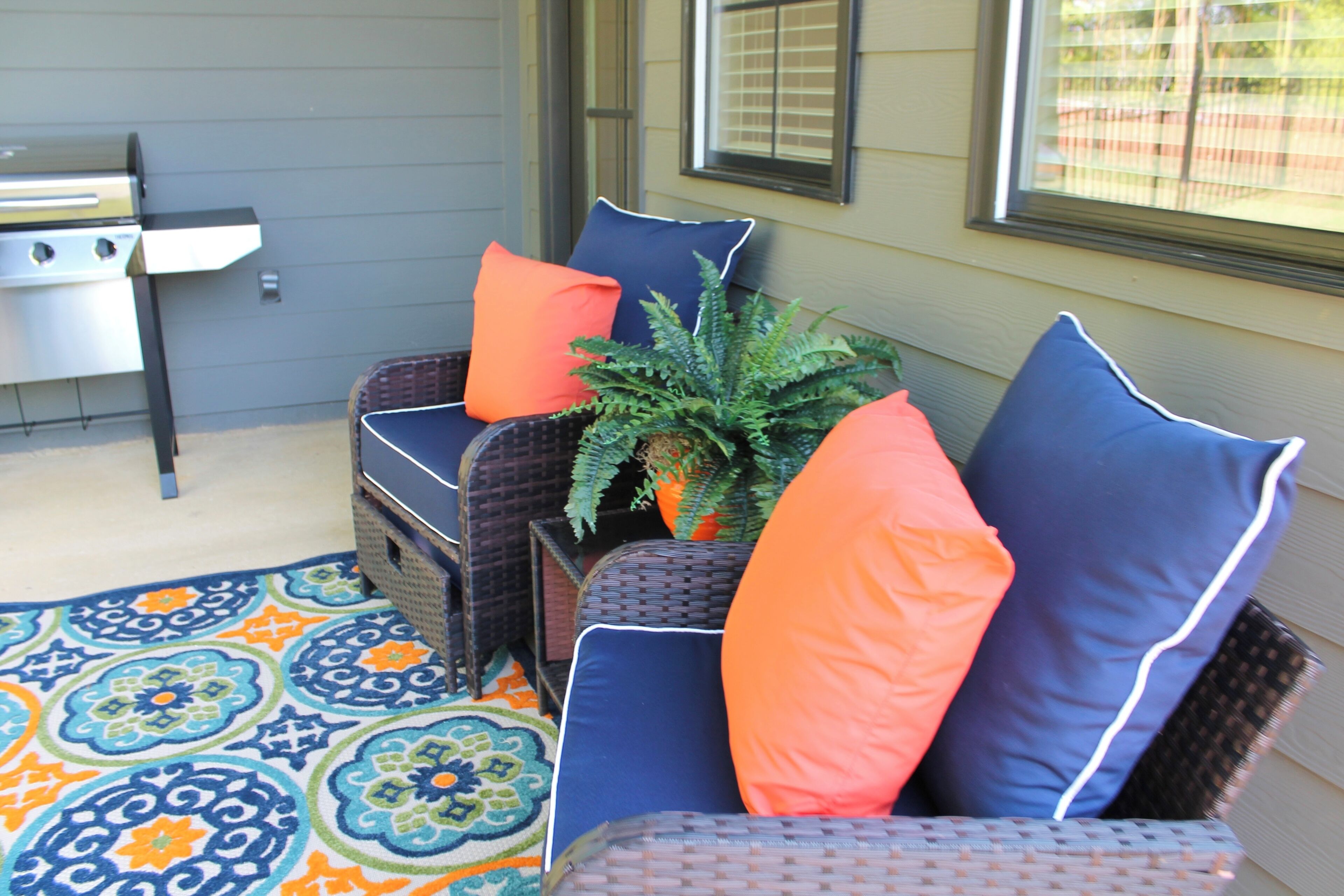 Un espace patio aménagé avec des meubles en osier, un tapis aux motifs colorés et un barbecue.