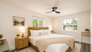A bedroom with a bed, nightstands, a ceiling fan, windows, and a painting on the wall.