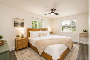 A bedroom with a bed, nightstands, a ceiling fan, windows, and a painting on the wall.