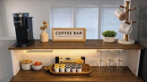 A coffee bar setup with a Keurig coffee maker, a sign reading 'Coffee Bar,' a small potted plant, a mug tree, and various coffee and tea containers.
