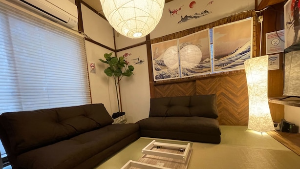 A living room with a dark brown sofa, a glass-top coffee table, a potted plant, and traditional Japanese wall art.