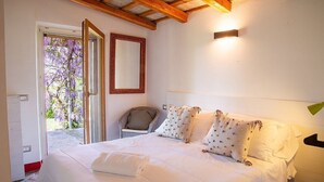 Un dormitorio con una cama grande, una silla, un espejo y una puerta que da a un área exterior con flores moradas.
