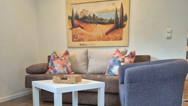 A living room with a brown sofa, two colorful pillows, a white coffee table, and a framed painting on the wall. - Vacation apartment with mountain view for max. 2 persons, 36 sqm (Bad Reichenhall)