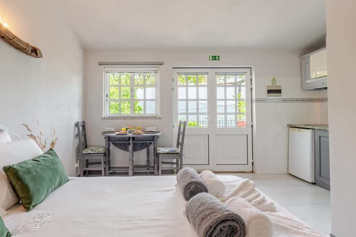 Quarto de hotel com uma cama, uma mesa de jantar com cadeiras, área de cozinha e vista para o verde lá fora.