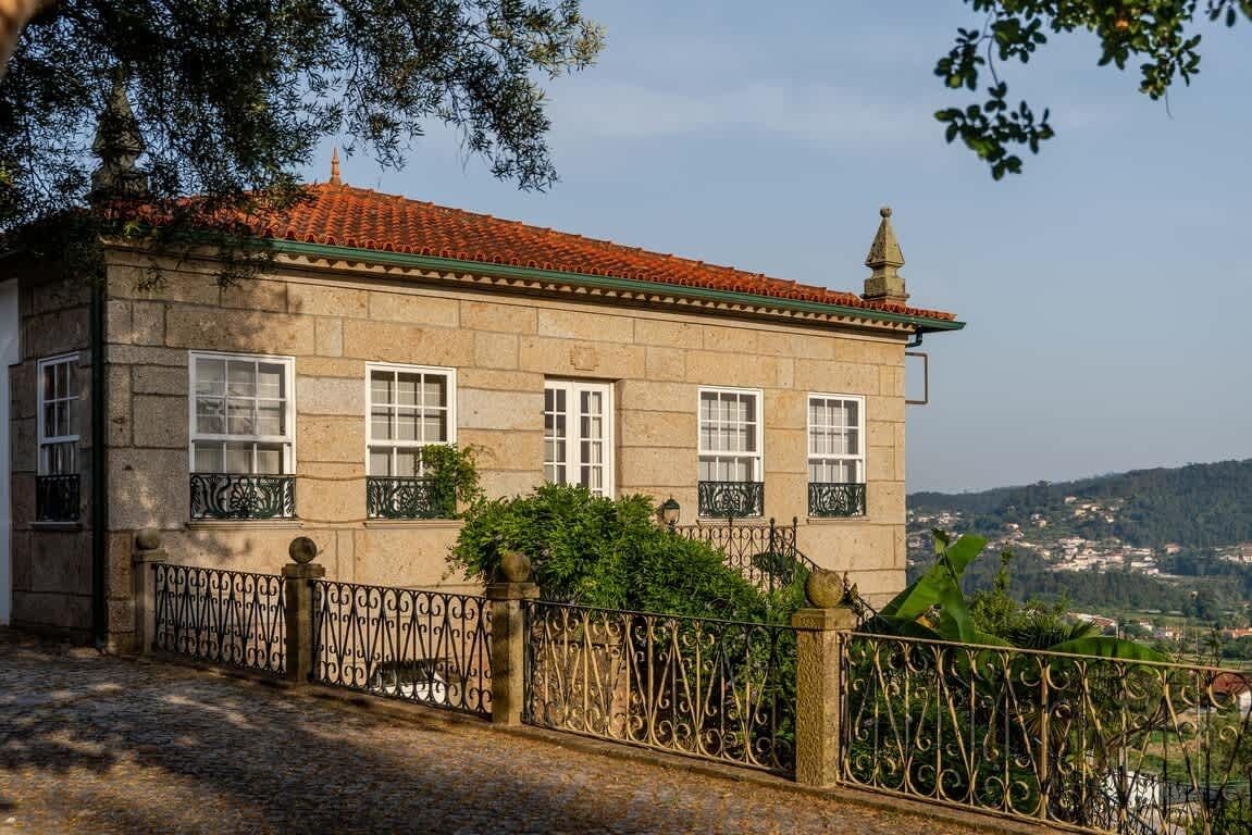 Un edificio de piedra con techo de tejas rojas, ventanas de marco blanco y una cerca de hierro forjado.
