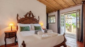 Una cama con un elaborada cabecera de madera, dos mesitas de noche con lámparas, y vista al exterior a través de una puerta abierta.
