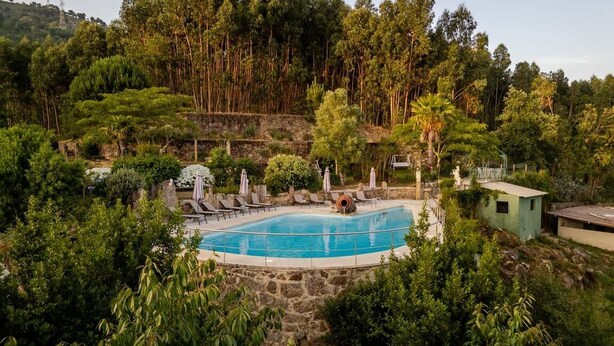 Una piscina rodeada de sillas de descanso y sombrillas, con un muro de piedra y vegetación al fondo.
