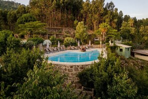 Una piscina rodeada de sillas de descanso y sombrillas, con un muro de piedra y vegetación al fondo.