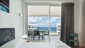Habitación de hotel con cama, escritorio y sillas, vista al océano y balcón con mesa y sillas.