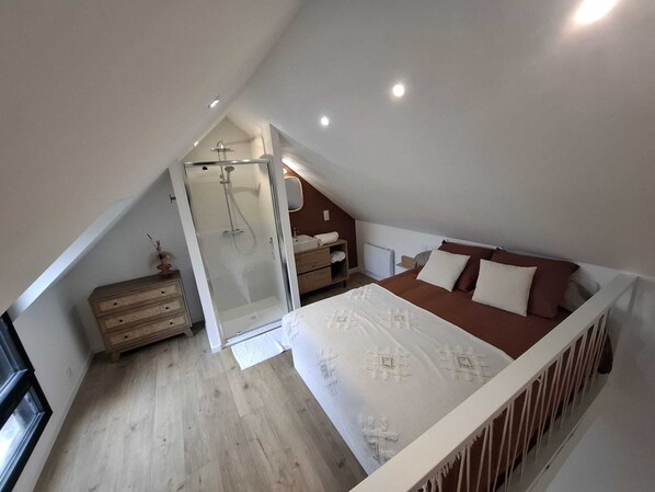 Une chambre avec un plafond en pente, un lit, une table de chevet, une commode, un dressing et une douche.