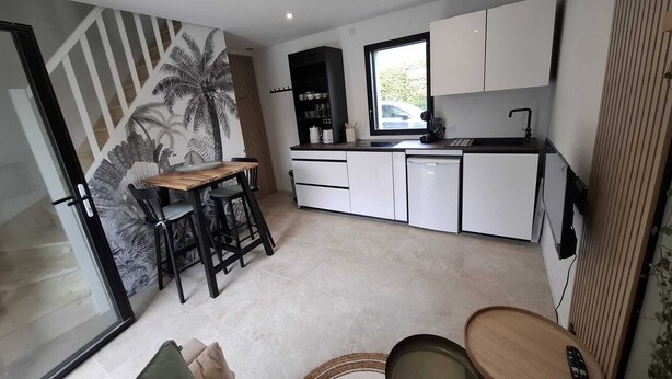 A compact kitchen with white cabinets, a black countertop, and a window. There is a dining area with a wooden table and two chairs. - Ty C&K Breizh (Plerguer)
