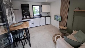 A compact kitchen with white cabinets, a black countertop, and a window. There is a dining area with a wooden table and chairs, and a small round table with a cushion. - Ty C&K Breizh (Plerguer)
