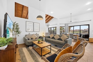 A modern living room with a sofa, coffee table, and wicker chairs. There is a TV on the left and a dining area with a view of the landscape.