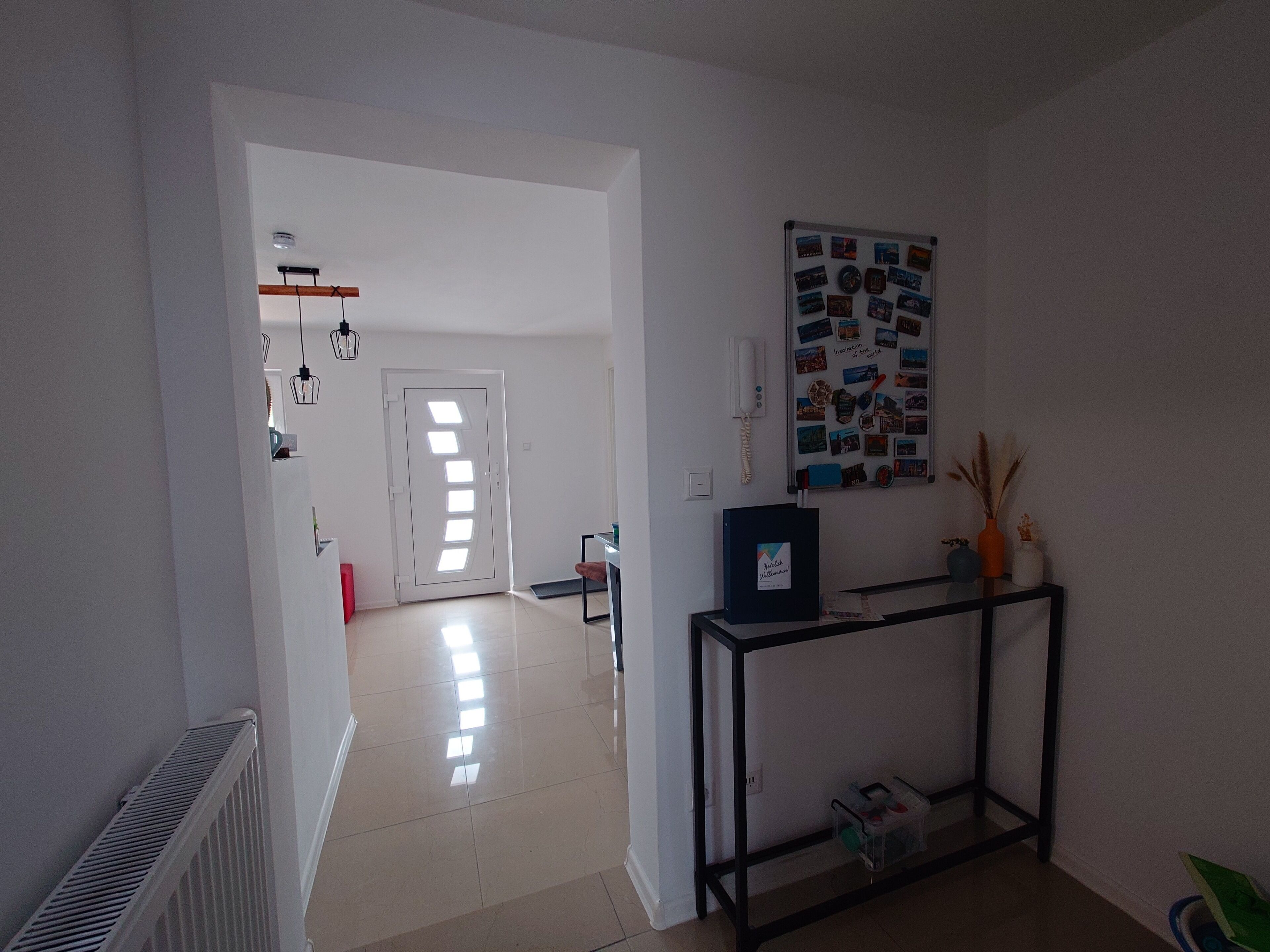 Un pasillo con una puerta blanca, un tablón de anuncios con fotografías y una pequeña mesa con marco negro.