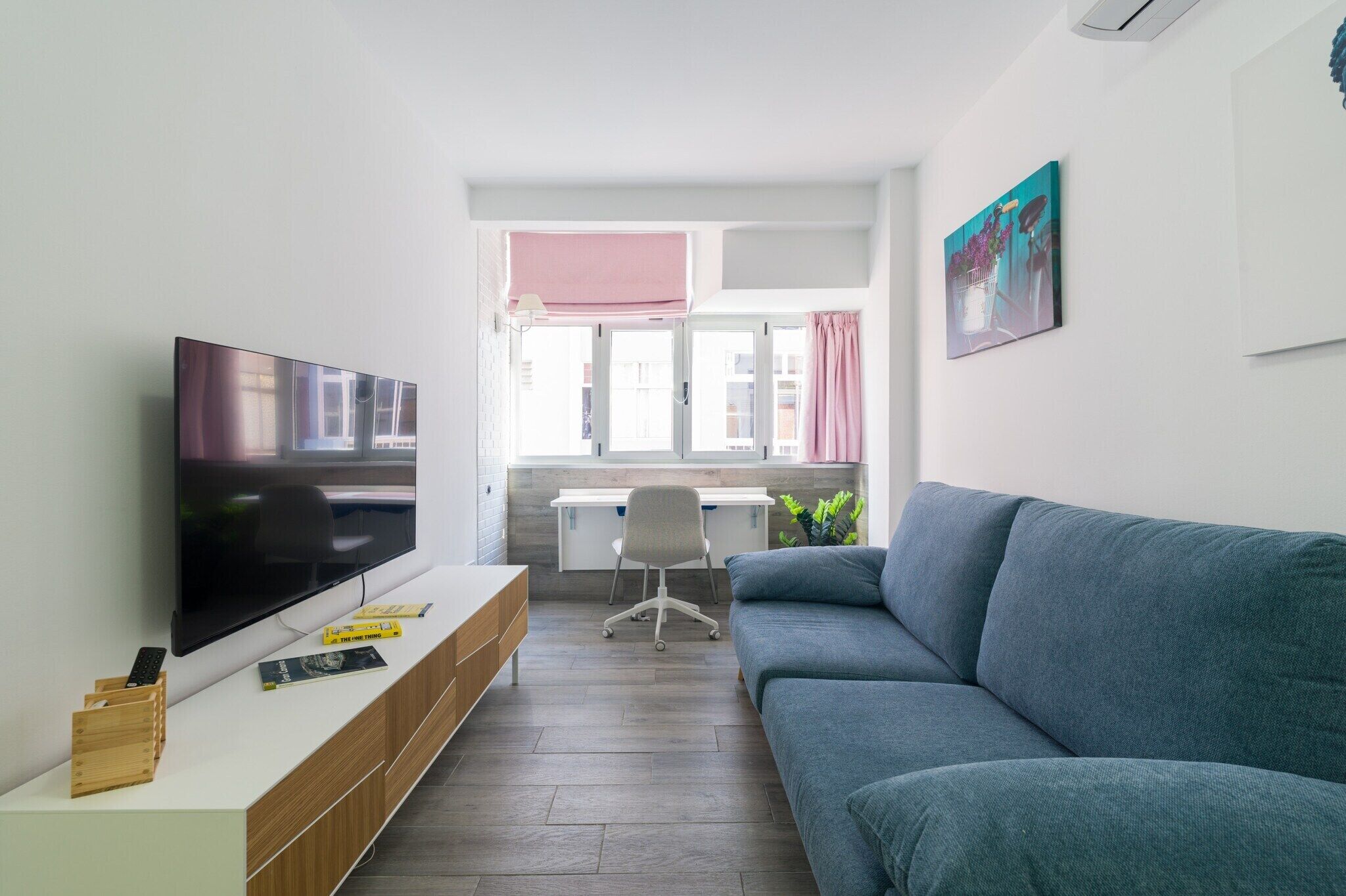 Un salon moderne avec un téléviseur à écran plat, un canapé gris, un meuble TV en bois, un bureau avec une chaise et une fenêtre avec des rideaux.