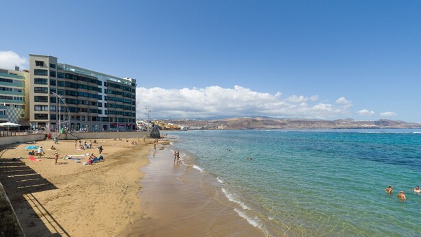 A beach with a hotel in the background, clear blue water, and people swimming.