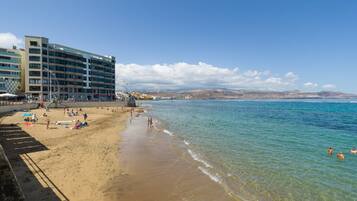 A beach with a hotel in the background, clear blue water, and people swimming.