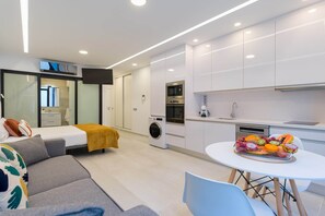 A modern hotel room with a bed, a sofa, a dining table with fruit, a washing machine, and a kitchen area.