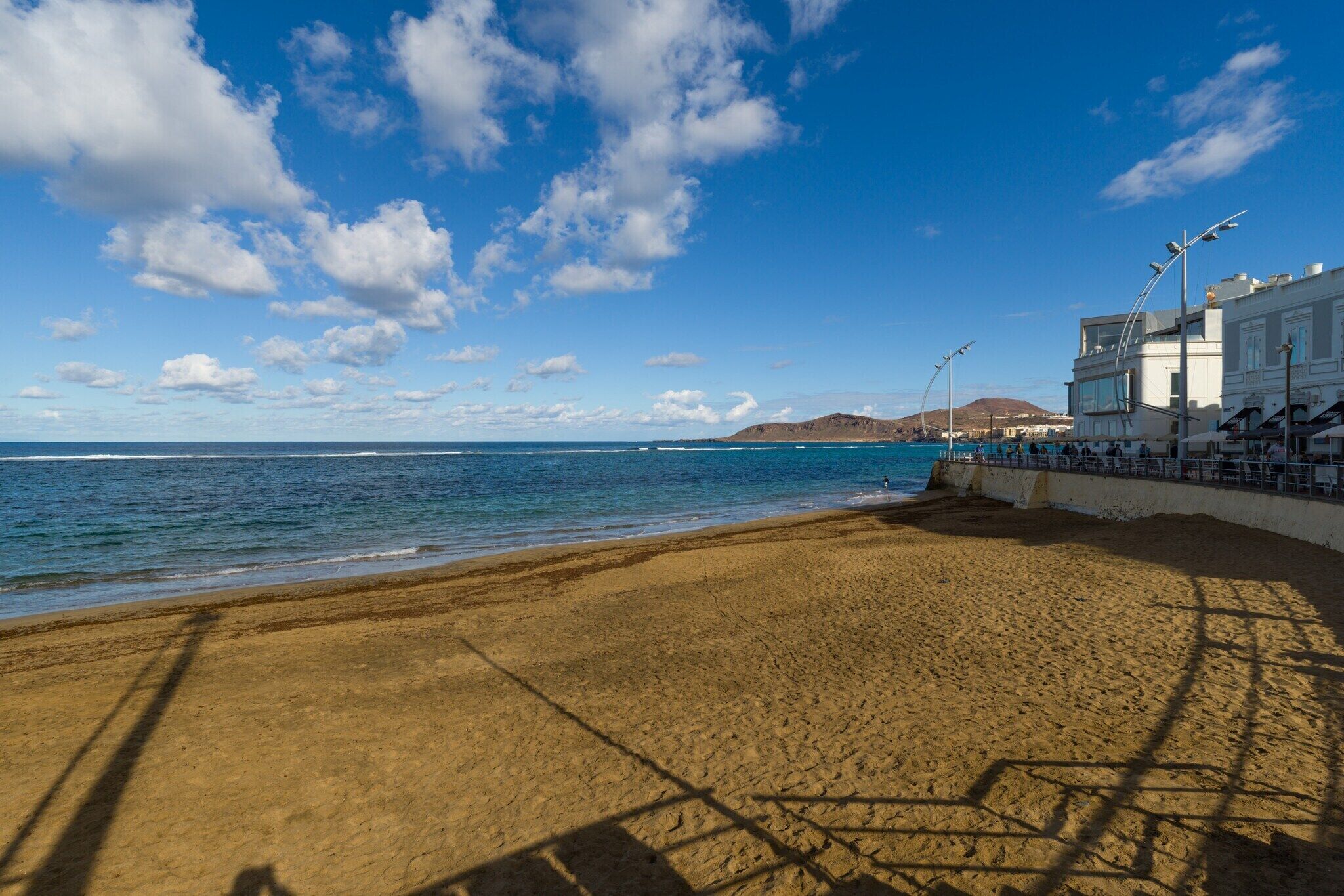 Ein Strand mit blauem Himmel und ruhiger See. Ein Gebäude mit Balkon ist rechts zu sehen.
