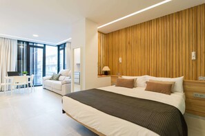A modern hotel room with a large bed, a desk with a laptop, a sofa, and a view of the city through large windows.