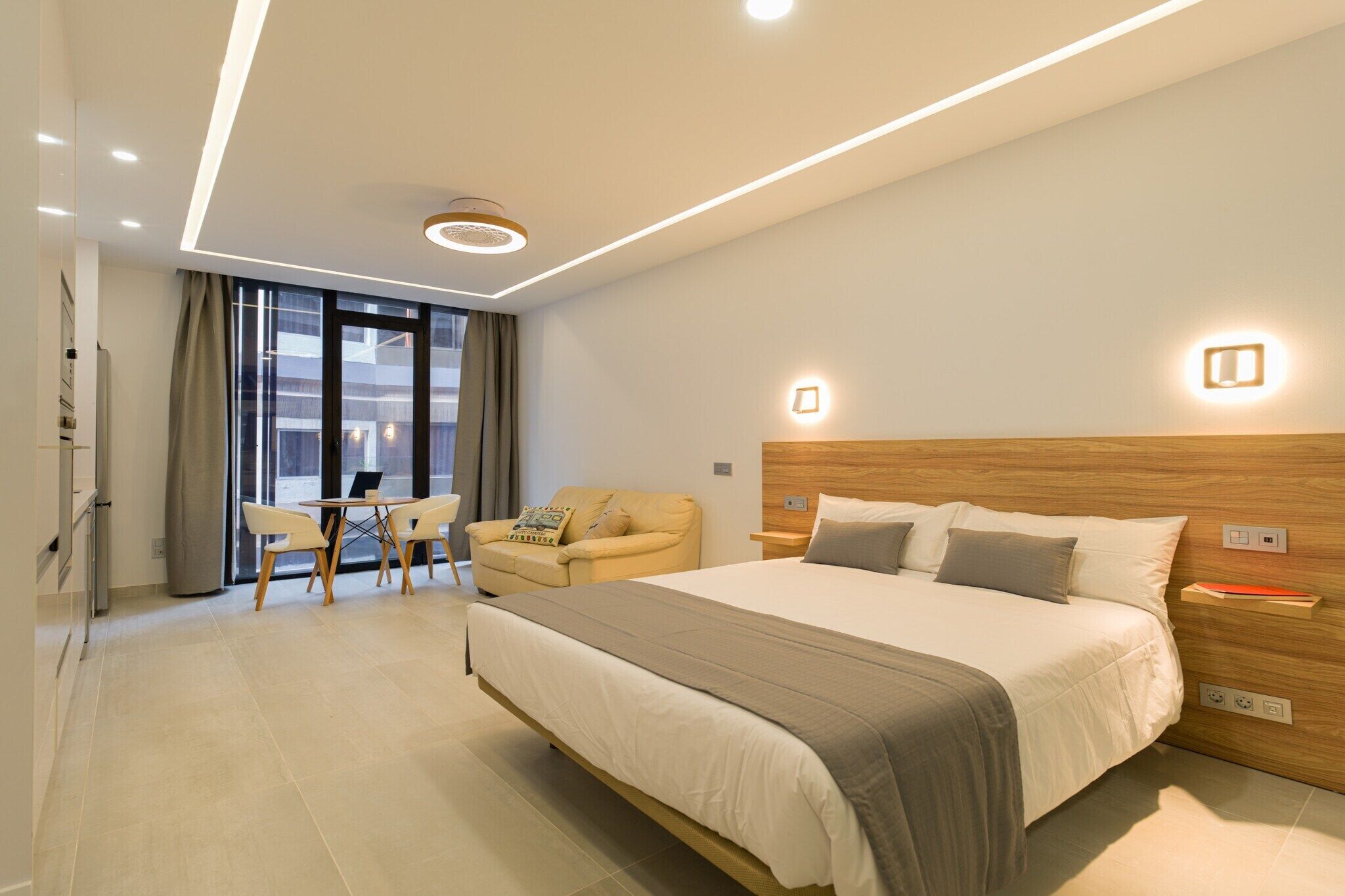 A modern hotel room with a large bed, a wooden headboard, a desk with a chair, and a sofa.