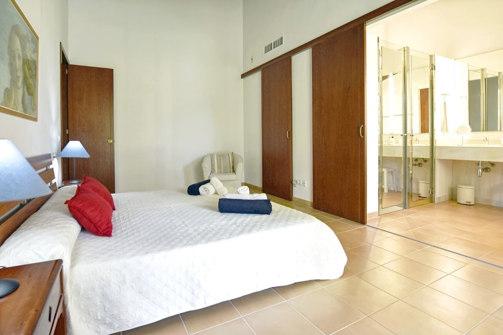 Een slaapkamer met een bed, een stoel, een bureau, een lamp, een kledingkast, en een badkamer met een spiegel en wastafel.