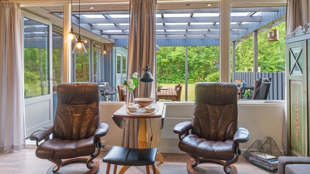 Une pièce chaleureuse avec deux fauteuils en cuir, une petite table et une vue sur un jardin grâce à de grandes fenêtres.