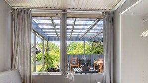 Une pièce avec un canapé gris, une petite table en bois sur laquelle se trouve une lampe, et un bureau blanc. Une grande fenêtre, ornée de rideaux, offre une vue sur un jardin et sur du mobilier d’extérieur.