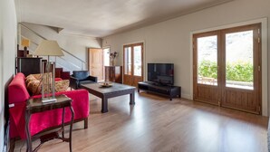 Un soggiorno ampio con un divano rosso, un tavolino, una TV e grandi porte in legno che si aprono su un balcone.