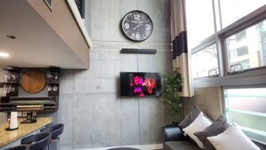Moderni oleskelutila, jossa on pyöreä musta rahi, tyynyillä varustettu sohva ja seinälle asennettu televisio.