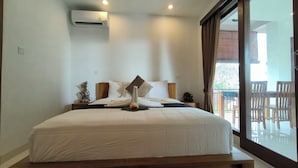 A neatly made bed with a white comforter and pillows, a bedside table with a decorative item, and a large window with a view of outdoor furniture. - Dharma Suweta (Ubud)