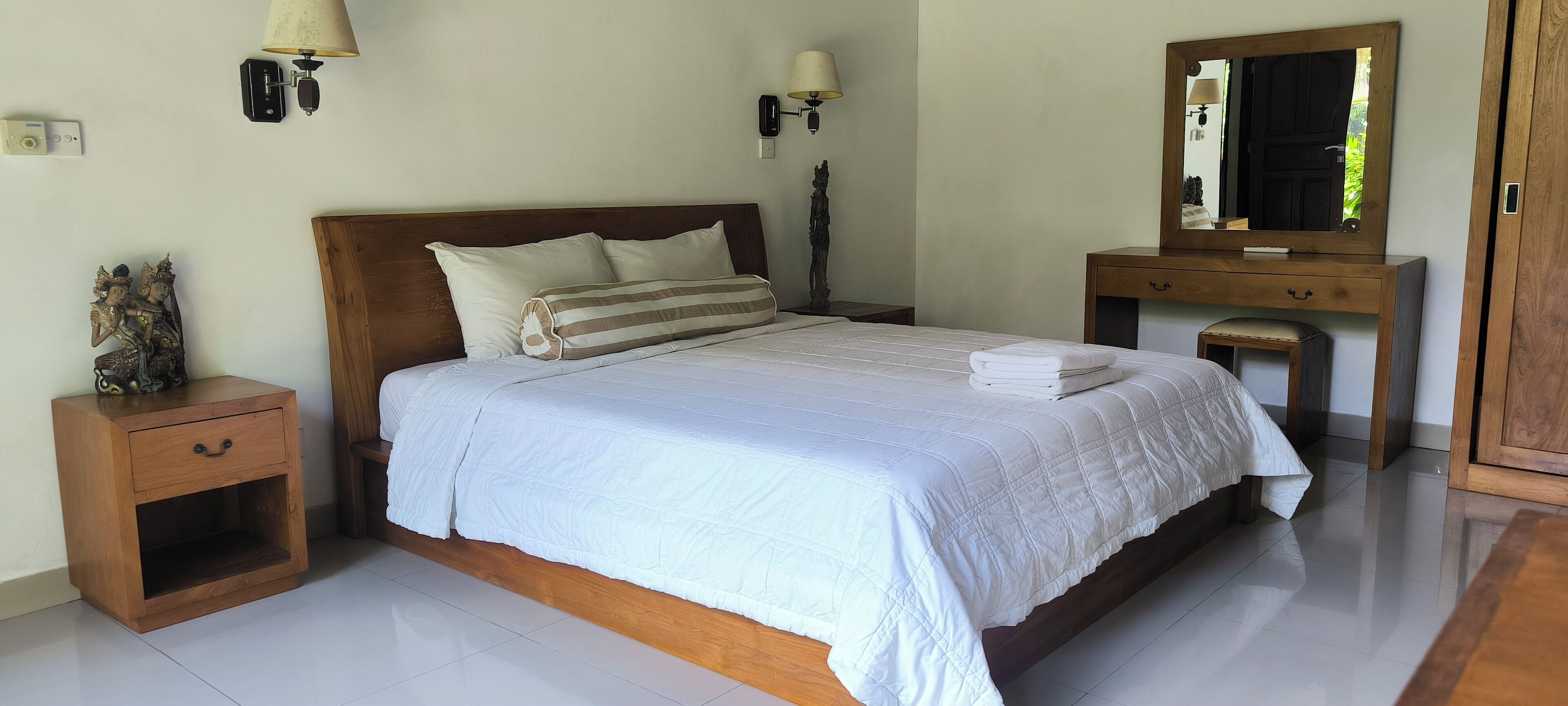 A neatly made bed with white linens and a striped pillow, a wooden nightstand with a drawer, a small table with a lamp, and a wooden dresser with a mirror.