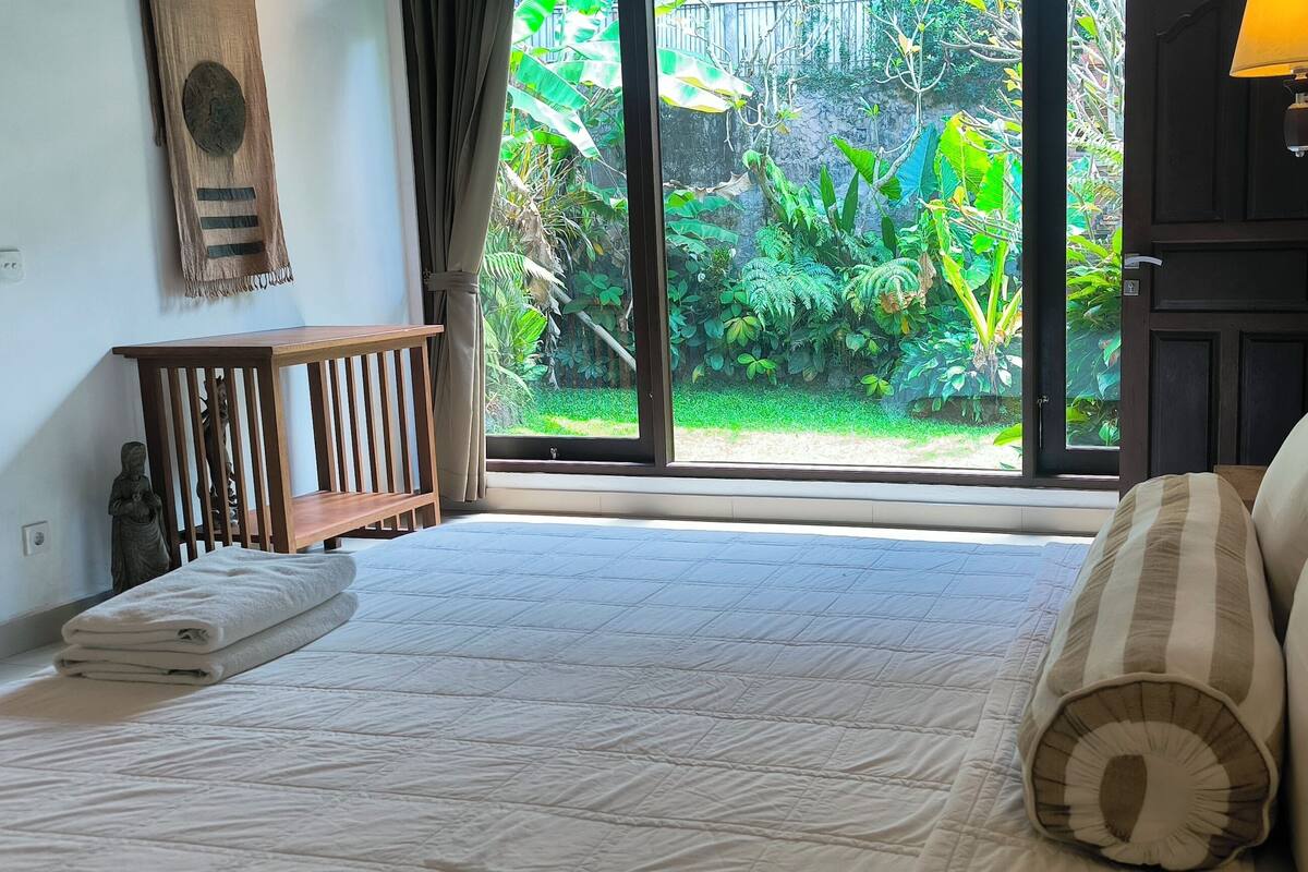 A bedroom with a large bed, a wooden nightstand, a lamp, and a view of a lush garden through a sliding door.