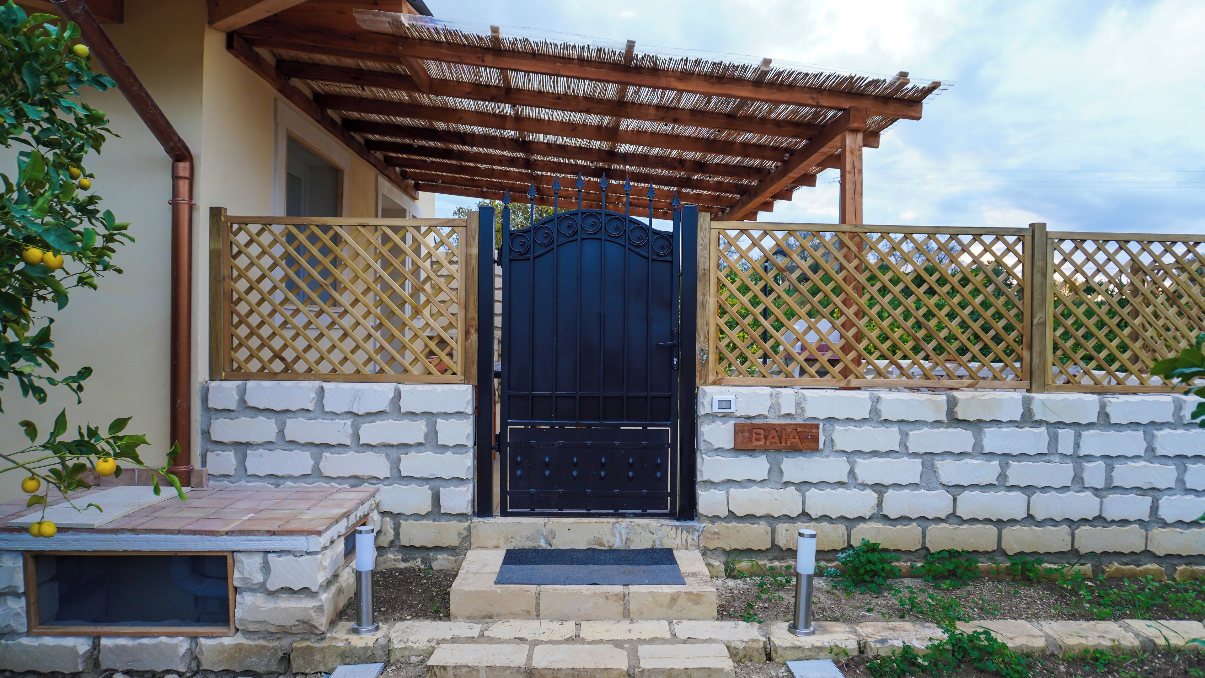 A black gate with a metal handle, a wooden nameplate reading "BAIA", and a stone wall with a tiled roof structure.