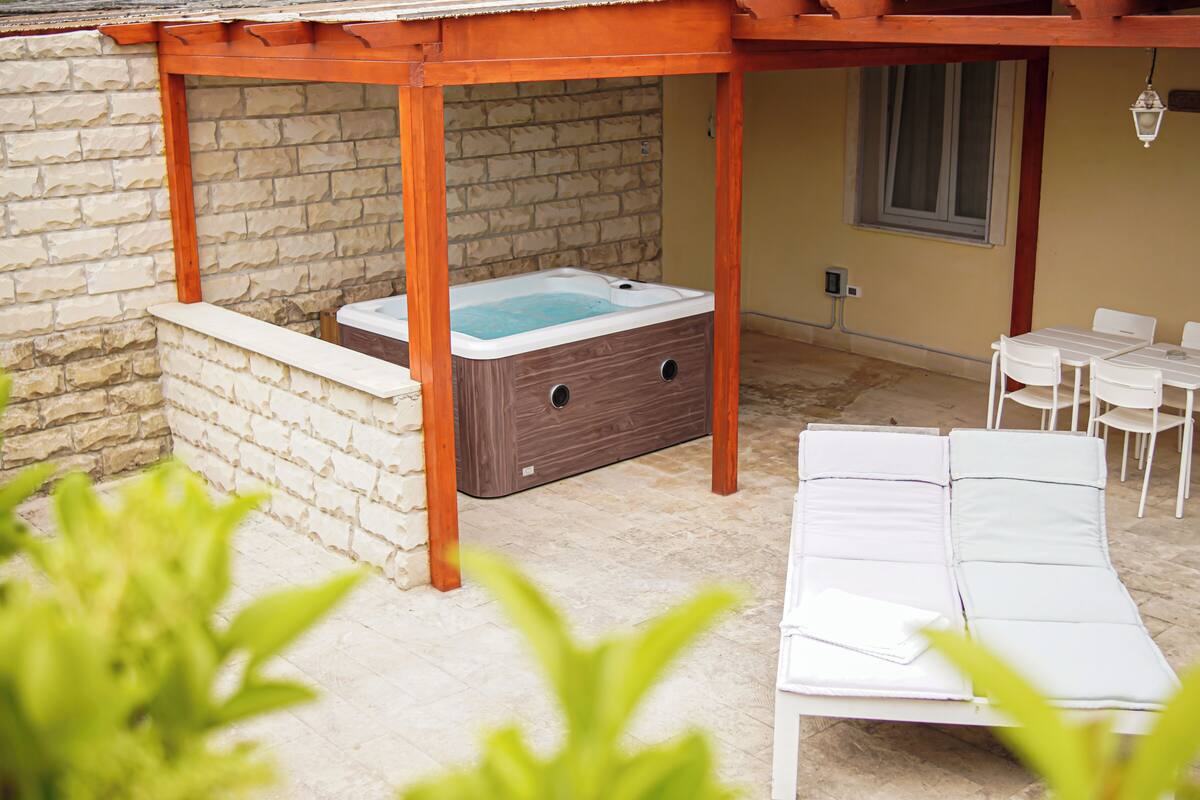 Un bain à remous sous une pergola en bois, un espace salon extérieur blanc et un mur en pierre.