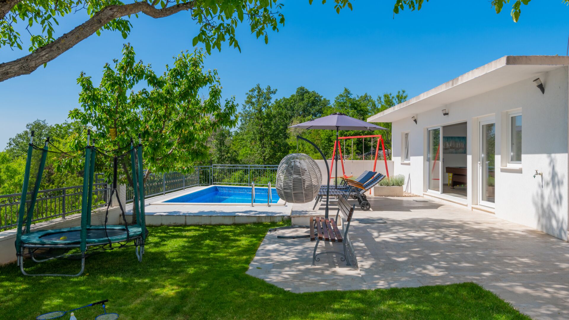 A backyard with a swimming pool, a swing set, and a patio area with outdoor furniture.