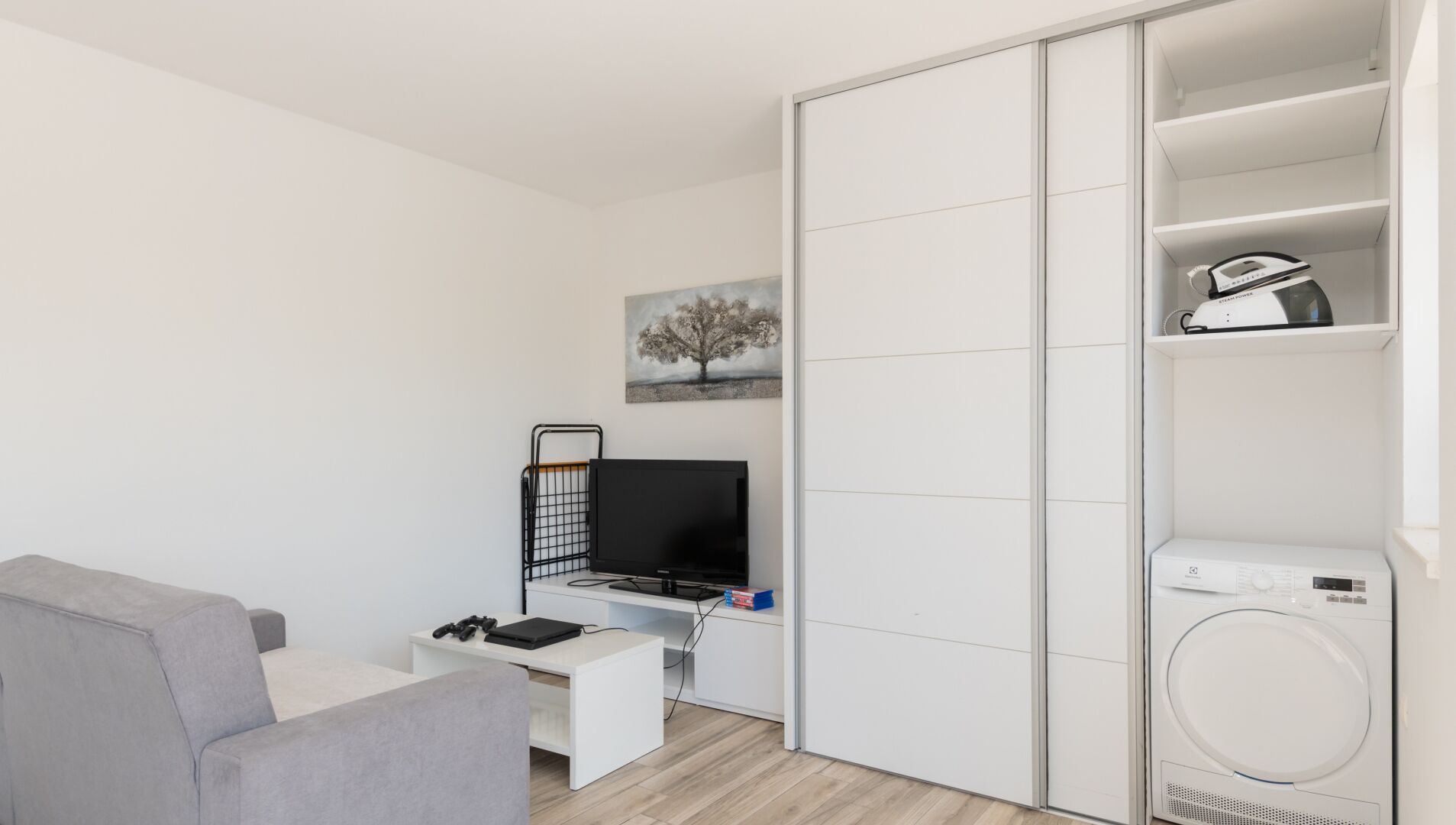 Un espacio habitable moderno y funcional con un sofá gris, un televisor sobre un mueble bajo, un escritorio con computadora y audífonos, un cuadro de un árbol y una lavadora.