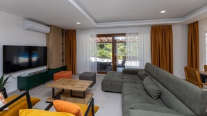 A modern living room with a large flat-screen TV, a grey sofa, a wooden coffee table, and orange and yellow cushions.