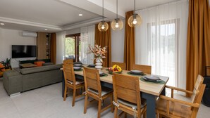 A modern dining area with wooden chairs and a table, a sofa, and a TV in the background.