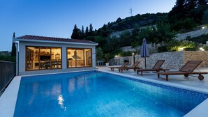 A swimming pool with lounge chairs, a glass-enclosed building with a tiled roof, and a stone wall with vegetation.