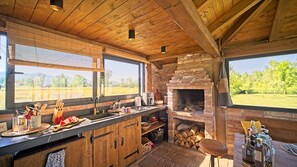 Uma cozinha rústica com lareira de tijolos, vigas de madeira e uma grande janela com vista para um campo.