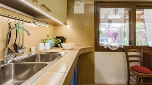 A kitchen with two stainless steel sinks, a wooden chair with a red cushion, and a window with a curtain. - Dimora Blu Poetto-Cagliari seaside Poetto beach (Sardegna)