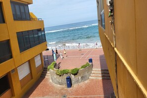 Un patio con vista al océano, con una jardinera circular con follaje, piso de ladrillo y un edificio amarillo con ventanas grandes.