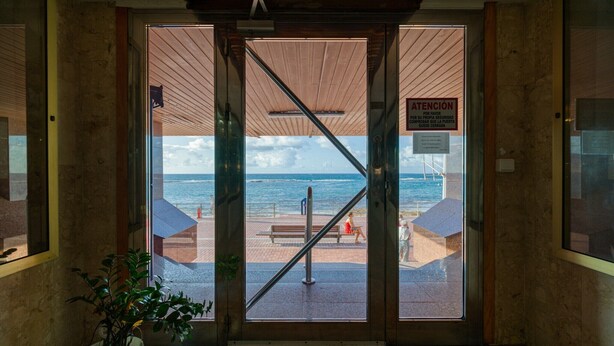 Vista a través de una puerta de vidrio con marco metálico, que da a una zona de playa con un banco y un letrero en español.
